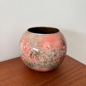 Peut inclure: Un vase décoratif rond avec une surface texturée. Le vase présente un mélange de teintes roses, rouges et grises, avec un intérieur brun foncé. Le récipient en céramique est posé sur une surface en bois.