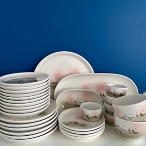 Puede incluir: Un juego de platos y cuencos de cerámica blanca con un diseño de ciudad rosa y verde. El juego incluye platos llanos, platos para ensalada, cuencos y una fuente para servir.