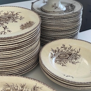 Op de afbeelding: Een grote collectie crèmekleurig keramisch servies met een bruin transferpatroon van een kalkoen omringd door gebladerte. De set bevat gestapelde borden, een schaal op voet en een ovale schaal, allemaal voorzien van een decoratieve rand.