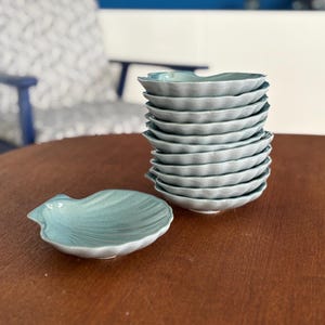 Peut inclure: Dix bols en céramique bleu clair en forme de coquillages empilés sur une table en bois.