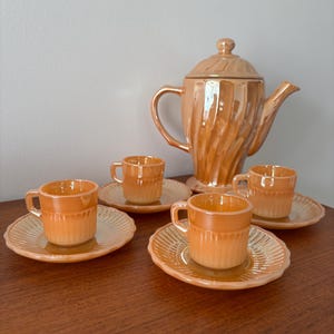 Könnte beinhalten: Ein Vintage-Teeservice in einem warmen Orangeton. Das Set beinhaltet eine Teekanne mit Deckel, vier Teetassen und vier Untertassen. Die Tassen und Untertassen haben ein geripptes Design. Die Teekanne hat einen gebogenen Griff und Ausguss.