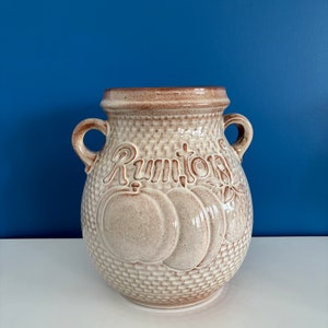 Könnte beinhalten: Eine weiße Keramik-Vase mit braunen Akzenten und einem gewebten Muster. Die Vase hat zwei Griffe und das Wort "Rumtopf" steht auf der Vorderseite.