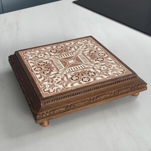 Peut inclure: Une boîte décorative carrée en bois avec un dessus en carrelage floral détaillé. Le carrelage présente un motif marron et crème. La boîte a un cadre en bois brun foncé et repose sur de petits pieds arrondis. La boîte est sur une surface blanche.