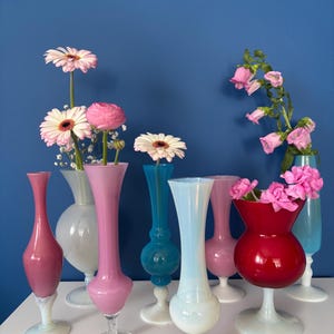 May include: A collection of colorful glass vases, each holding fresh flowers. The vases vary in shape and color, including pink, red, blue, and white. The flowers are a mix of pink and white daisies and other blooms, set against a blue background.