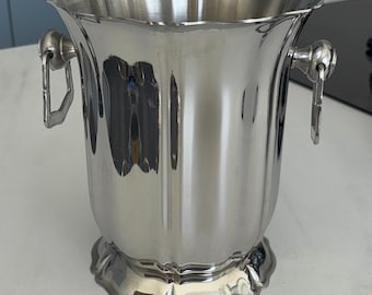 May include: A polished silver-tone metal ice bucket featuring a scalloped rim and base with vertical fluted detailing. The bucket includes two side-mounted ring handles attached to circular hardware, providing a reflective and metallic finish.
