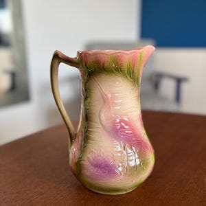 May include: A pink and green ceramic pitcher with a raised design of a bird and flowers. The pitcher has a handle and a wide mouth.