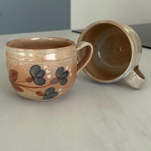 Peut inclure: Deux tasses en céramique avec des anses. L'une est droite, l'autre est sur le côté. Les tasses sont beiges avec un motif floral bleu, marron et blanc. Les tasses ont une texture mouchetée.