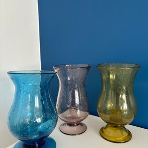 Op de afbeelding: Drie glazen vazen in tinten blauw, paars en geelgroen. Elke vaas heeft een bolvormig lichaam met een uitlopende rand en een cirkelvormige basis. De vazen worden tentoongesteld op een wit oppervlak tegen een blauw-witte achtergrond.