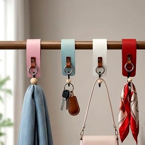 Könnte beinhalten: Vier lederartige Aufhängeschlaufen in Rosa, Hellblau, Weiß und Rot, jeweils mit einer braunen Lederlasche, einem silbernen Metallring und einer kleinen Holzperle. Die Schlaufen hängen an einer horizontalen Holzstange und halten einen blauen Schal, Schlüssel, eine Handtasche und einen gemusterten roten Schal.