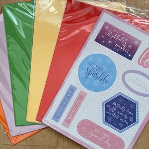 May include: A selection of coloured craft paper sheets in orange, lilac, green, yellow, and red. A sheet of stickers with phrases such as "Birthday wishes," "Let your day sparkle," and "With love" is also included. The stickers have a pale pink border.