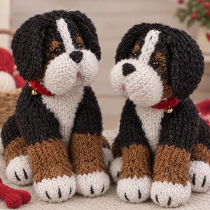 May include: Two handmade knitted plush dogs featuring black, brown, and white yarn. Each dog has a white stripe on the face, black ears, brown legs, and white paws. They wear red collars with small gold bells around their necks and have dark eyes.
