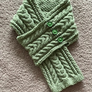 Könnte beinhalten: Ein grüner Strick Schal mit einem Zopfmuster und drei grünen Knöpfen.