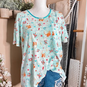 May include: A turquoise blue short-sleeved shirt with a pattern of cats, birds, and geometric shapes. The shirt has a turquoise blue trim around the neckline and a small pocket on the left side.