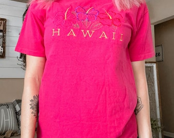 May include: A bright pink t-shirt featuring embroidered hibiscus flowers and the word "HAWAII" in gold lettering. The shirt has short sleeves and a crew neck. The design includes pink, purple, and yellow floral details.