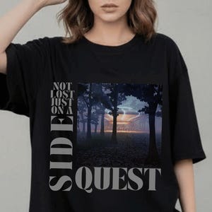 Könnte beinhalten: Schwarzes übergroßes T-Shirt mit Landschaftsgrafik und dem Text "NOT LOST JUST ON A SIDE QUEST". Die Grafik zeigt eine Waldszene mit Sonnenuntergang. Das Shirt wird mit hellblauen Jeans kombiniert.