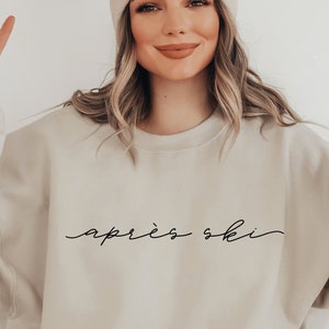 Könnte beinhalten: Ein beiger Pullover mit dem Schriftzug "après ski" in schwarzer Kursivschrift.