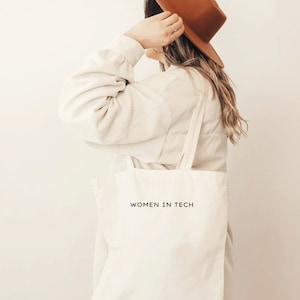 Op de afbeelding: Een persoon draagt een bruine hoed en een wit shirt en houdt een witte tote bag vast met de tekst "WOMEN IN TECH" erop gedrukt.