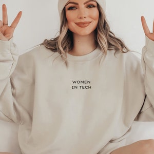 Könnte beinhalten: Ein beigefarbenes Sweatshirt mit Rundhalsausschnitt und dem schwarzen Schriftzug "WOMEN IN TECH" mittig auf der Brust, kombiniert mit einer passenden beigen Strickmütze. Das Kleidungsstück hat lange Ärmel mit gerippten Bündchen und einen lockeren Schnitt.