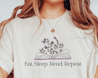 Peut inclure: Un t-shirt à manches courtes de couleur crème arborant un motif de livre ouvert avec des fleurs violettes sortant des pages. Sous l'illustration, le texte "Eat. Sleep. Read. Repeat." est écrit en police serif noire. Le t-shirt a un col rond.