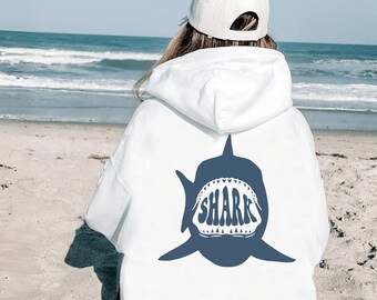 May include: A white hooded sweatshirt featuring a large dark blue graphic on the back. The graphic depicts a shark silhouette with an open mouth containing the word SHARK in bold, stylised lettering. The hoodie includes a hood and long sleeves.