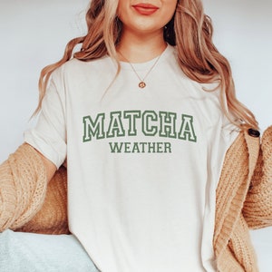 May include: A white t-shirt with the text "MATCHA WEATHER" in green lettering. The shirt is being worn by a person with long blonde hair and a brown cardigan.