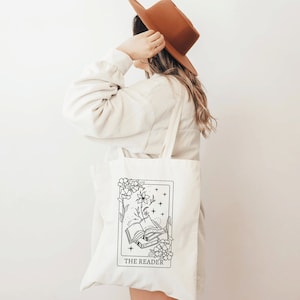 Op de afbeelding: Een witte canvas tote bag met een zwart-witte illustratie van een tarotkaart. De kaart toont een open boek met een bloem die eruit groeit. De tekst "THE READER" is onder de kaart gedrukt.