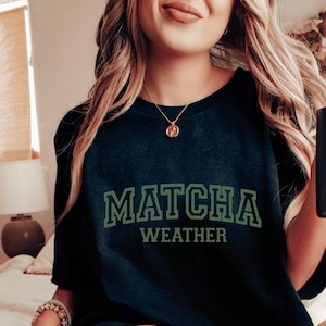 May include: A black t-shirt with the text "MATCHA WEATHER" in green letters. The shirt is worn by a person with long blonde hair.