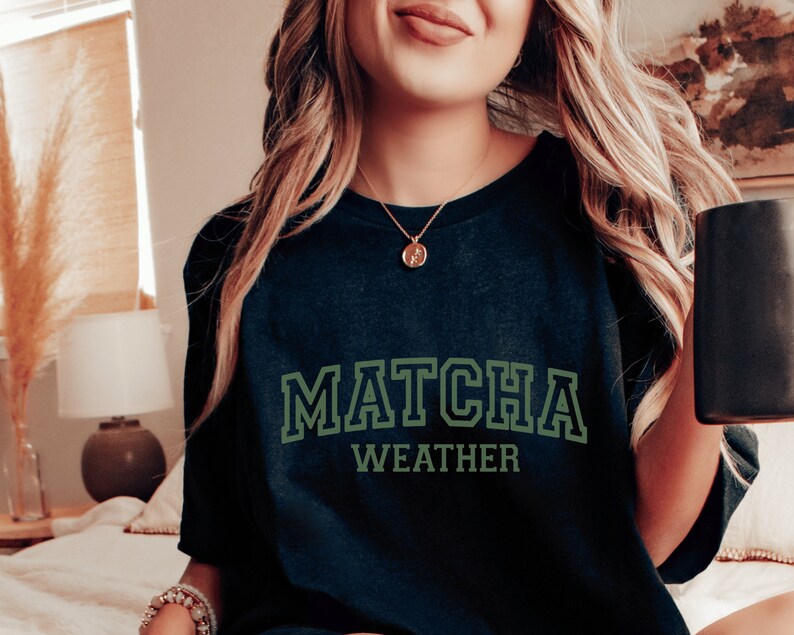 May include: A black t-shirt with the text "MATCHA WEATHER" in green letters. The shirt is worn by a person with long blonde hair.