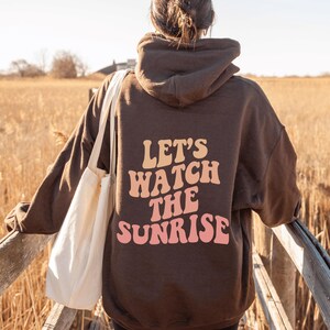 May include: A brown hoodie with a white tote bag. The hoodie has the text "Let's Watch The Sunrise" printed on the back in pink and orange letters.