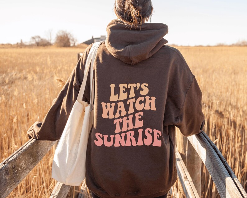 May include: A brown hoodie with a white tote bag. The hoodie has the text "Let's Watch The Sunrise" printed on the back in pink and orange letters.