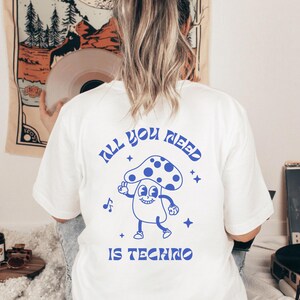 Puede incluir: Camiseta blanca con un dise&ntilde;o azul en la espalda que dice "All You Need Is Techno" con un dise&ntilde;o de hongo de dibujos animados. La camiseta es de estilo casual.