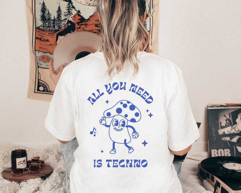 Puede incluir: Camiseta blanca con un dise&ntilde;o azul en la espalda que dice "All You Need Is Techno" con un dise&ntilde;o de hongo de dibujos animados. La camiseta es de estilo casual.
