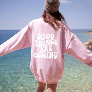 Könnte beinhalten: Rosafarbener Pullover mit dem Text "GOOD THINGS ARE COMING" in weißer Retro-Schrift.