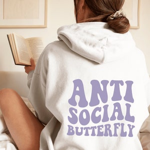 May include: A light gray hoodie with the words "ANTI SOCIAL BUTTERFLY" in lavender lettering on the back. The hoodie has a hood and long sleeves. The person is reading a book.