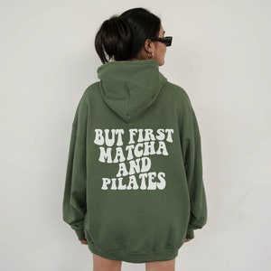 May include: An olive green hooded sweatshirt featuring the white text BUT FIRST MATCHA AND PILATES printed on the back. The garment has long sleeves, a hood, and a relaxed fit, with the bold, wavy-style lettering centred on the back panel.