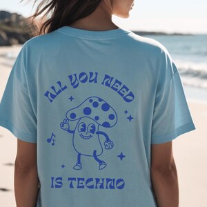 Puede incluir: Camiseta azul claro con un dise&ntilde;o retro en la espalda. El dise&ntilde;o presenta un personaje de dibujos animados de un hongo con el texto "ALL YOU NEED IS TECHNO" en azul. La camiseta es de corte holgado.