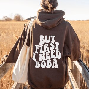 Może przedstawiać: Brązowa bluza z kapturem z nadrukiem "BUT FIRST I NEED BOBA" na plecach w białych retro literach. Osoba nosi bluzę i trzyma białą torbę.