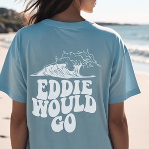 May include: A light blue t-shirt with a white graphic of a wave and the words "EDDIE WOULD GO" in a bold, retro font. The shirt is made of a soft material and is perfect for casual wear.
