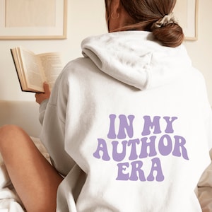 Könnte beinhalten: Ein weißer Hoodie mit einem lilafarbenen Aufdruck "In My Author Era" auf der Rückseite. Die Person trägt den Hoodie und liest ein Buch.
