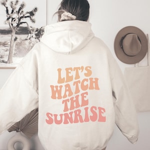May include: A cream-colored hoodie with a large graphic print on the back that says "Let's Watch The Sunrise" in a retro font with orange and pink colors.