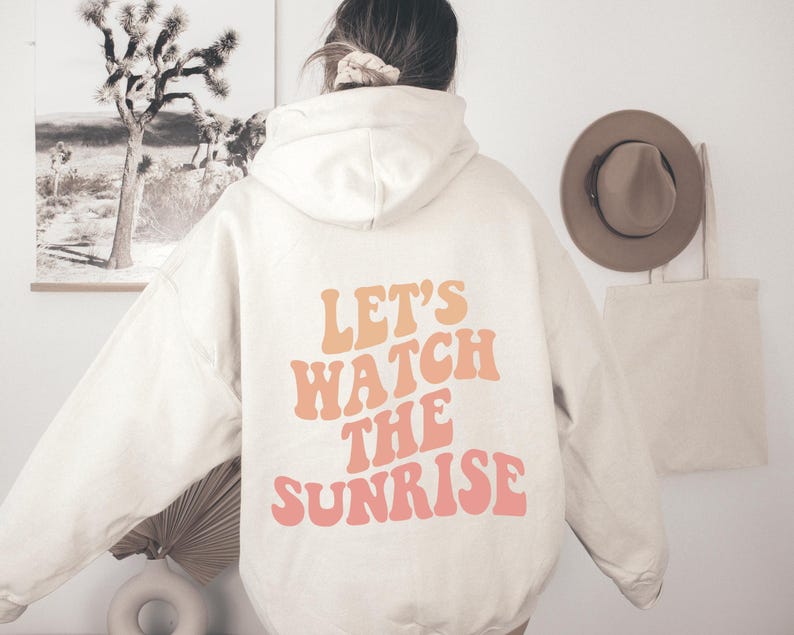May include: A cream-colored hoodie with a large graphic print on the back that says "Let's Watch The Sunrise" in a retro font with orange and pink colors.