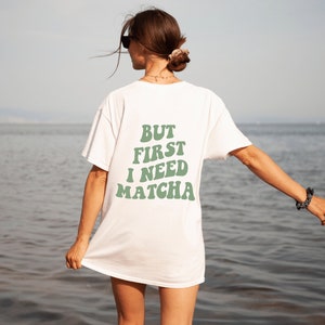 Könnte beinhalten: Weißes T-Shirt mit dem Text "BUT FIRST I NEED MATCHA" in Grün auf der Rückseite.