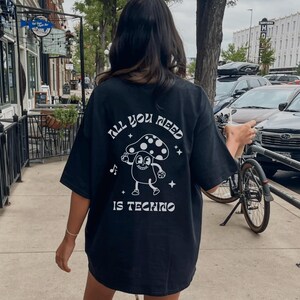 Puede incluir: Camiseta negra con el texto "ALL YOU NEED IS TECHNO" y un personaje de dibujos animados de un hongo. El hongo tiene un gorro blanco con lunares y est&aacute; bailando. La camiseta es holgada.