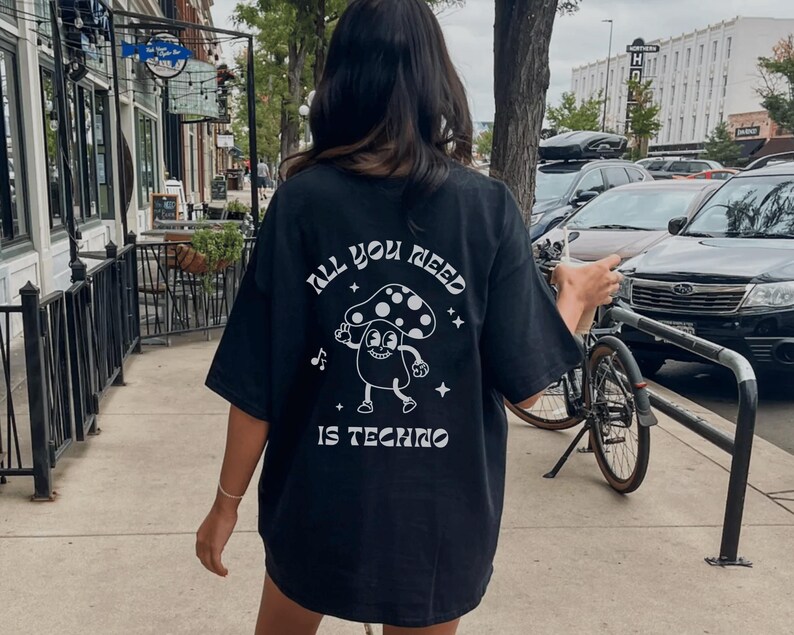 Puede incluir: Camiseta negra con el texto "ALL YOU NEED IS TECHNO" y un personaje de dibujos animados de un hongo. El hongo tiene un gorro blanco con lunares y est&aacute; bailando. La camiseta es holgada.
