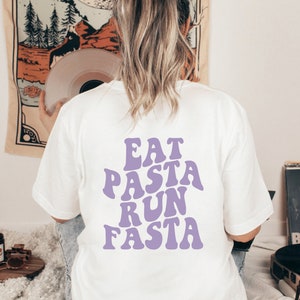 Könnte beinhalten: Ein weißes kurzärmeliges T-Shirt mit dem violetten Schriftzug EAT PASTA RUN FASTA, der in einer wellenförmigen, fetten Schrift auf dem Rücken gedruckt ist. Das Shirt hat einen Rundhalsausschnitt und eine Standardpassform mit dem mittigen Aufdruck.