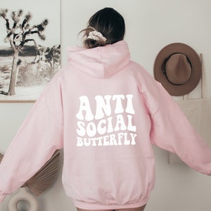 May include: A light pink hoodie with the words "ANTI SOCIAL BUTTERFLY" in white, retro-style lettering across the back. The hoodie has a hood and long sleeves. The image is taken from the back, showing the design on the garment.