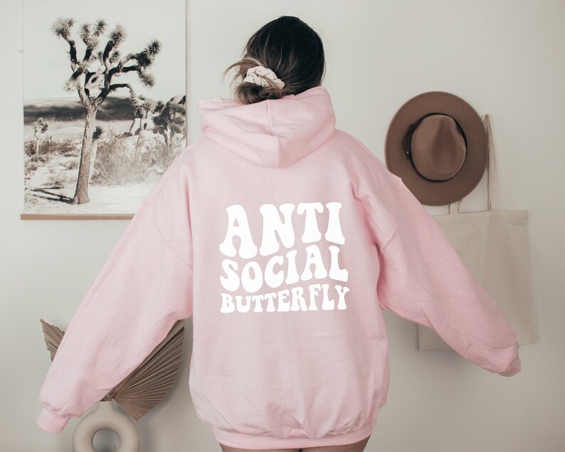 May include: A light pink hoodie with the words "ANTI SOCIAL BUTTERFLY" in white, retro-style lettering across the back. The hoodie has a hood and long sleeves. The image is taken from the back, showing the design on the garment.