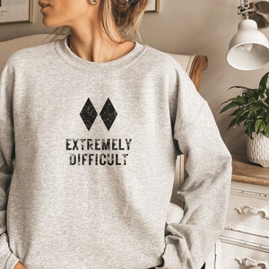 May include: A light gray sweatshirt with the words "EXTREMELY DIFFICULT" printed in black, below two black diamond shapes. The sweatshirt is a crew neck style and appears to be made of a soft material.