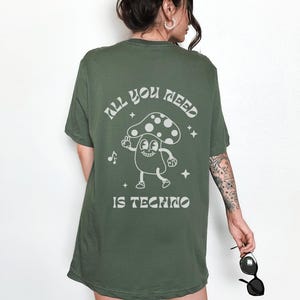 Puede incluir: Camiseta verde oliva con un gr&aacute;fico de hongo y el texto "ALL YOU NEED IS TECHNO". El dise&ntilde;o presenta un personaje de hongo de dibujos animados, notas musicales y estrellas.