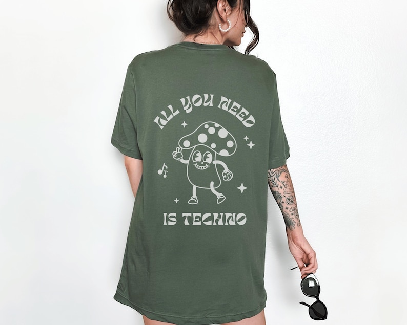 Puede incluir: Camiseta verde oliva con un gr&aacute;fico de hongo y el texto "ALL YOU NEED IS TECHNO". El dise&ntilde;o presenta un personaje de hongo de dibujos animados, notas musicales y estrellas.